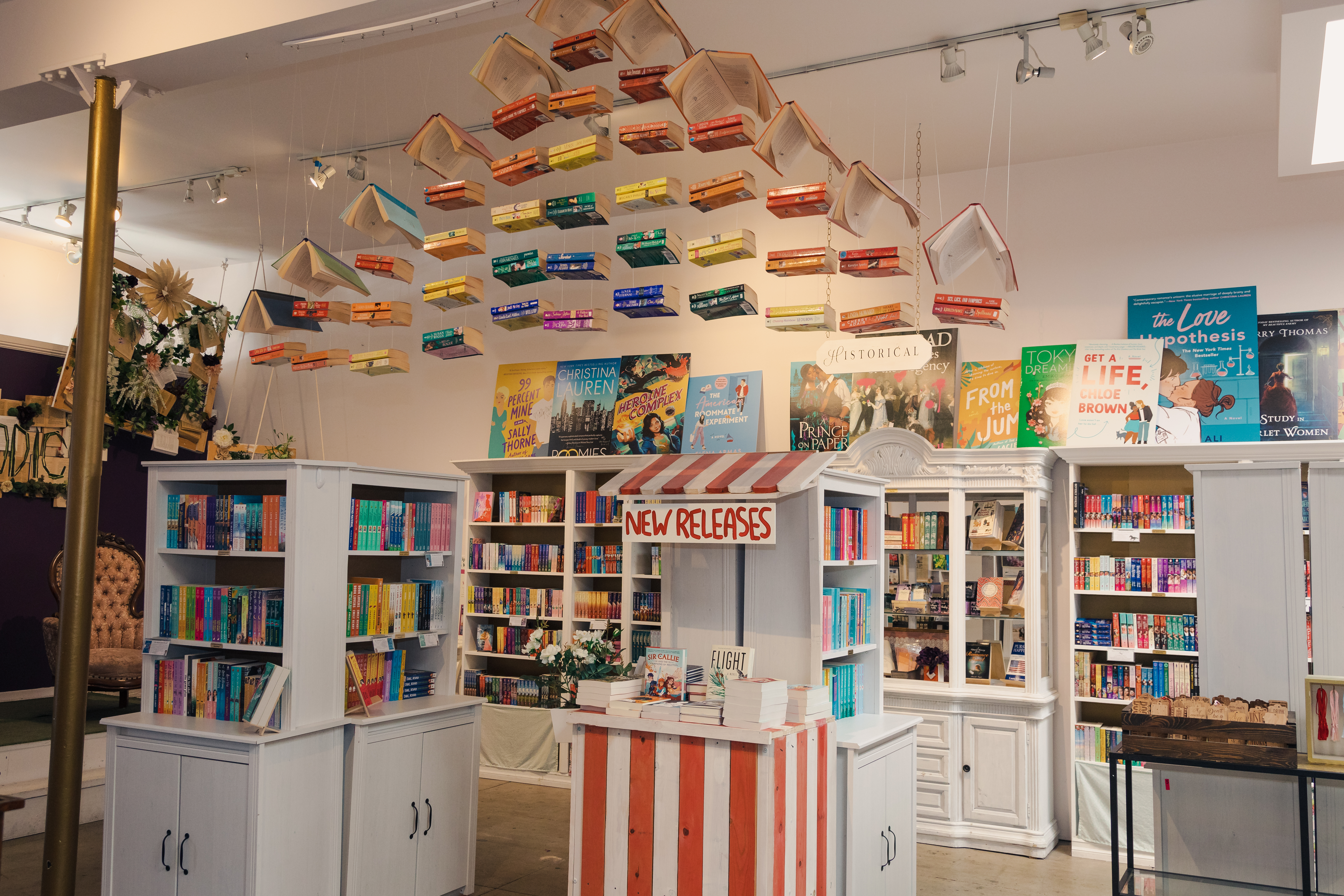 An image of the interior of The Ripped Bodice bookstore in Los Angeles. There’s a book cart that reads “new releases” and a hanging installation showing off a rainbow of book spines.