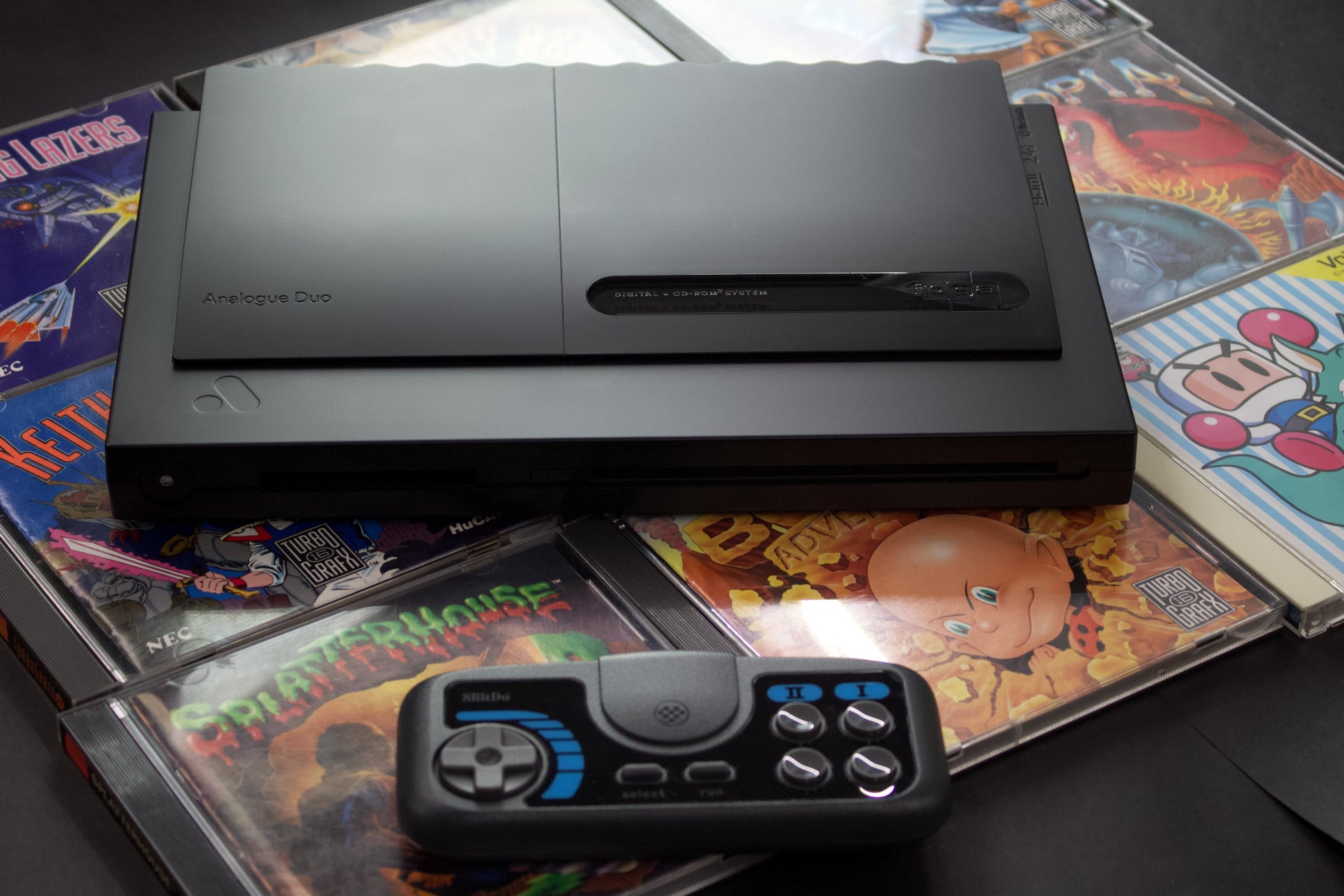 The Analogue Duo, on top of several game CD cases, and a blue and black controller beneath it