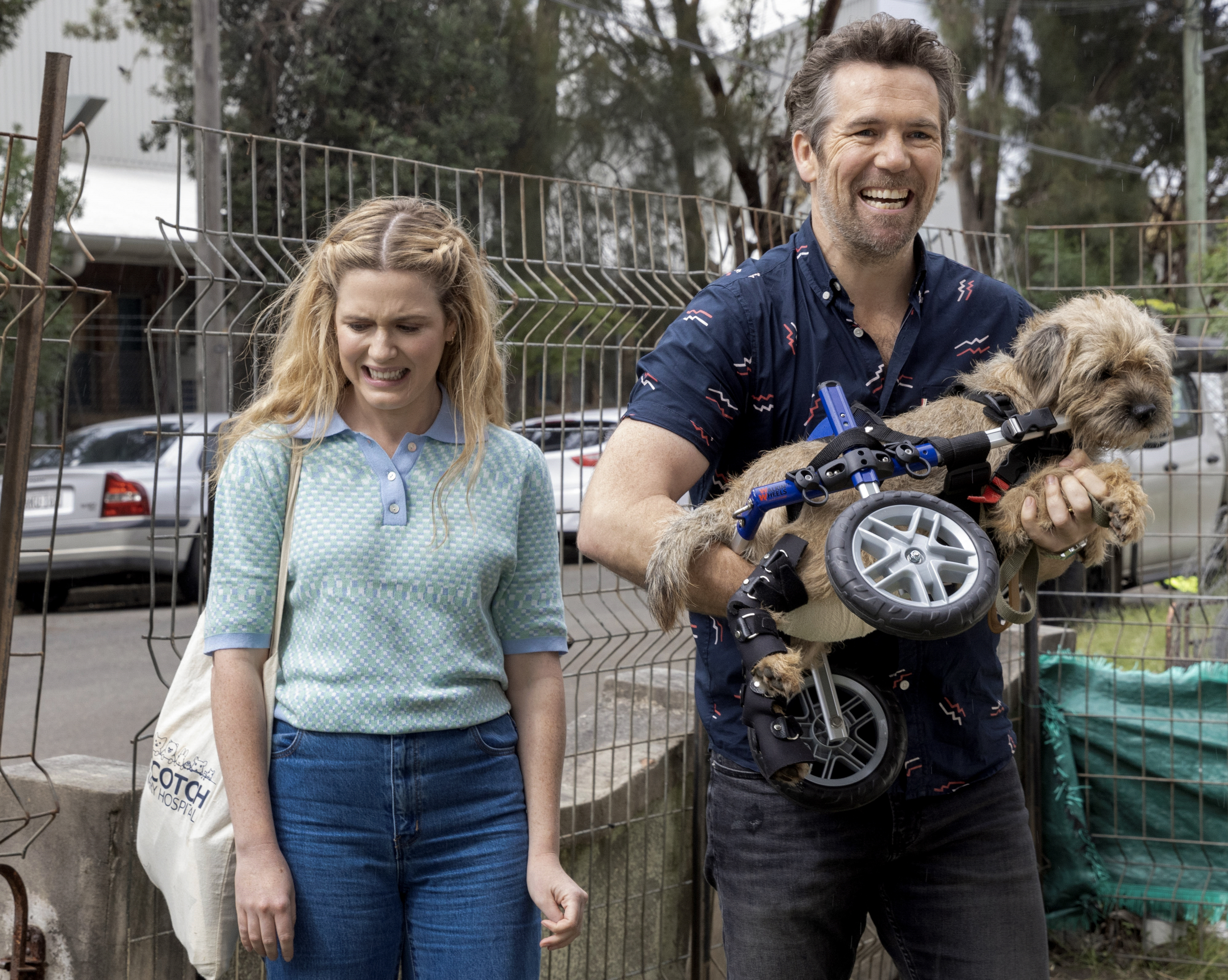 Ashley, a young woman with blonde hair, pulls an awkward face, while Gordon, a good looking middle aged guy, grins and carries a dog with wheels for back legs, in Colin From Accounts