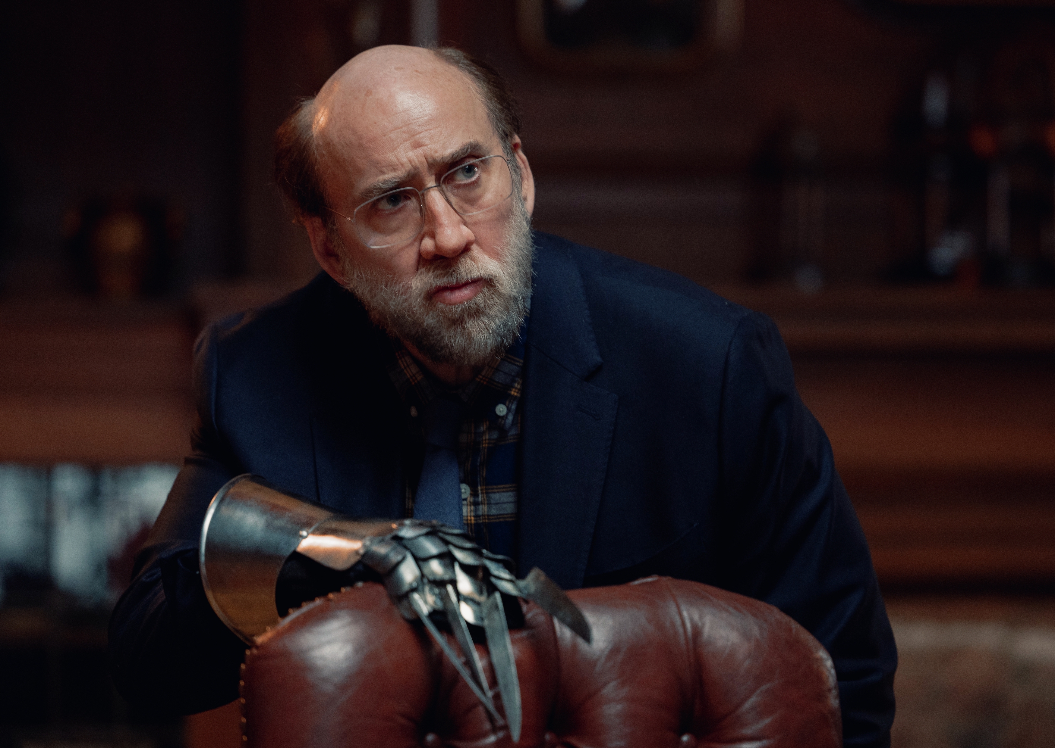 Schlubby professor Paul Matthews (Nicolas Cage) stands in a dark-paneled study in front of a big leather chair, posing with a clawed metal glove vaguely reminiscent of Freddy Krueger’s in a scene from A24’s Dream Scenario