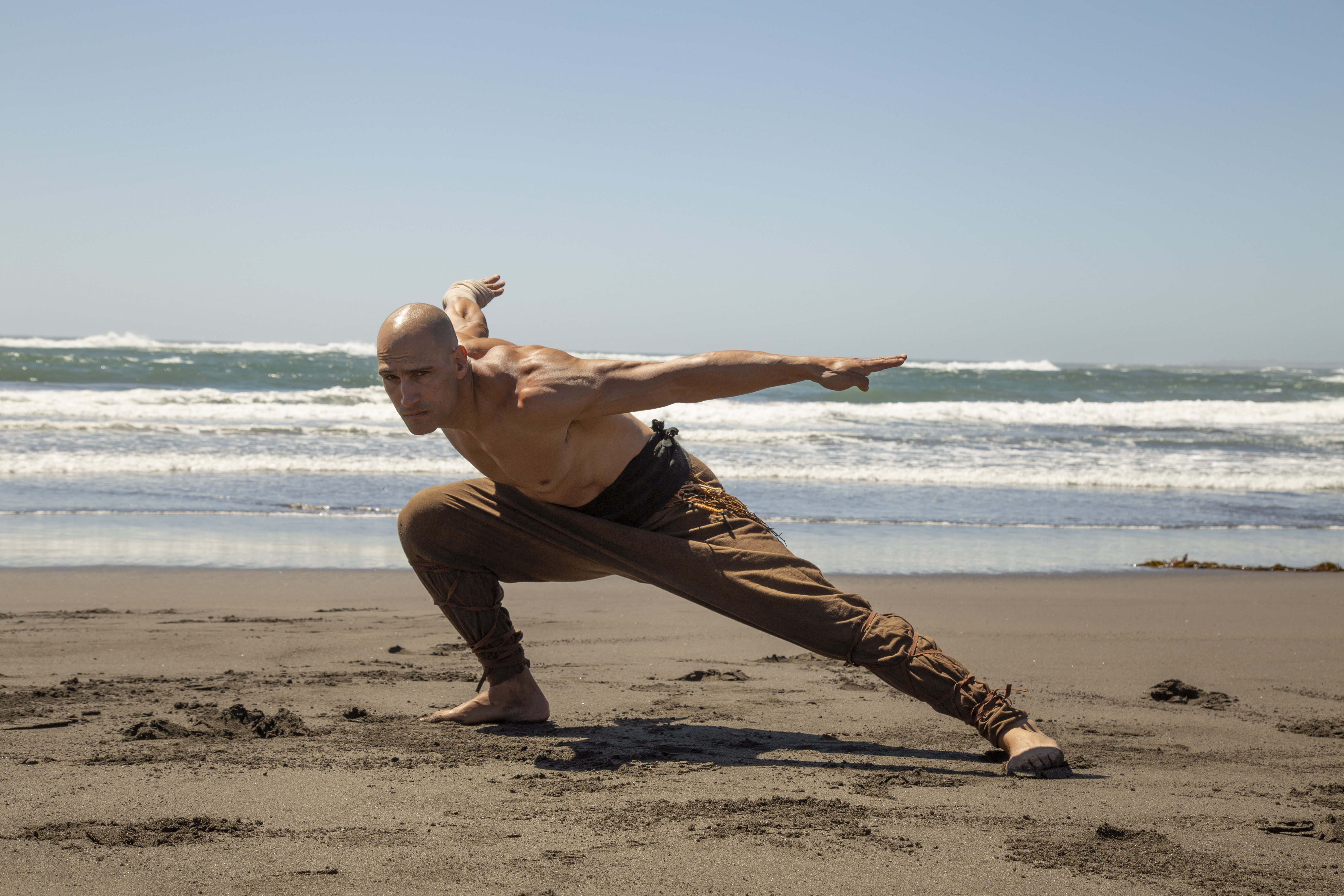 Marko Zaror crouches low in a defensive pose on a beach in Fist of the Condor, with the ocean behind him.