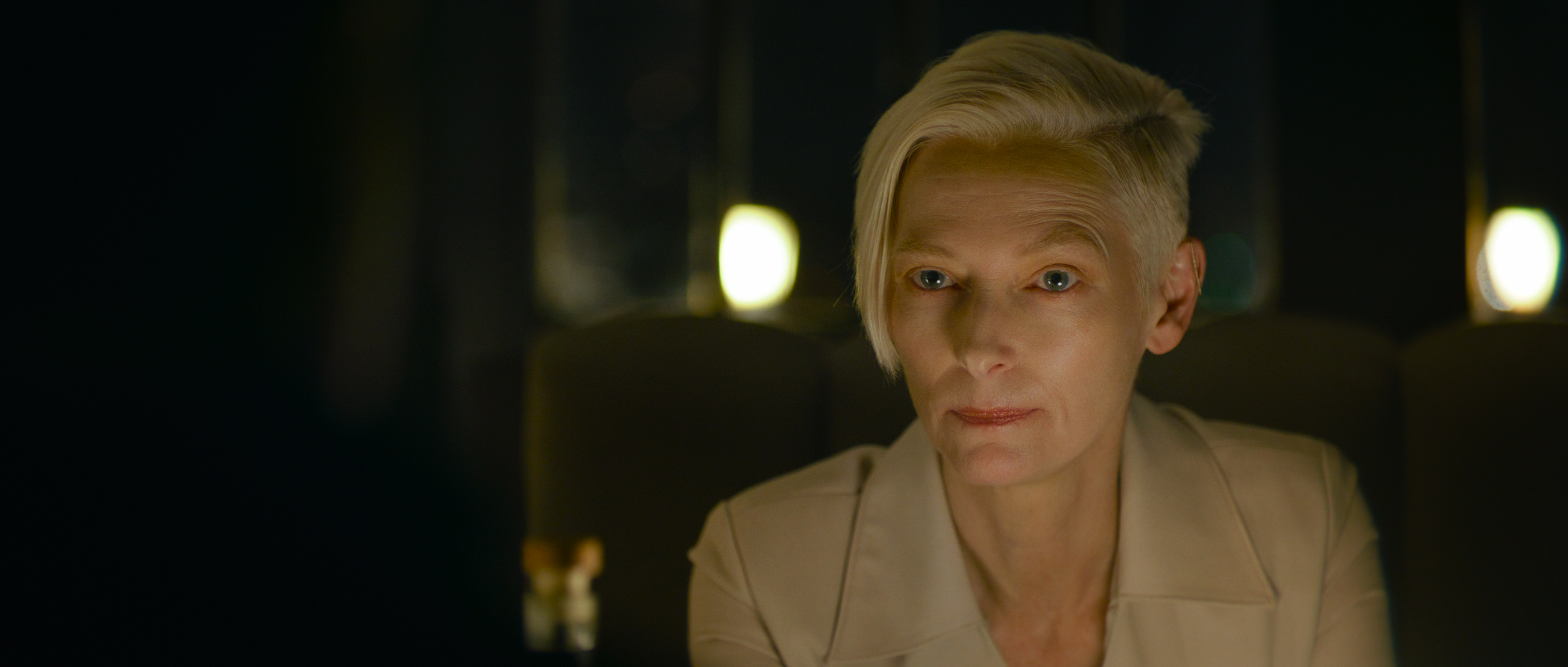 Tilda Swinton, with swooped-over white hair and wearing a white collared shirt, sits in the dark and looks intently into the camera in David Fincher’s The Killer