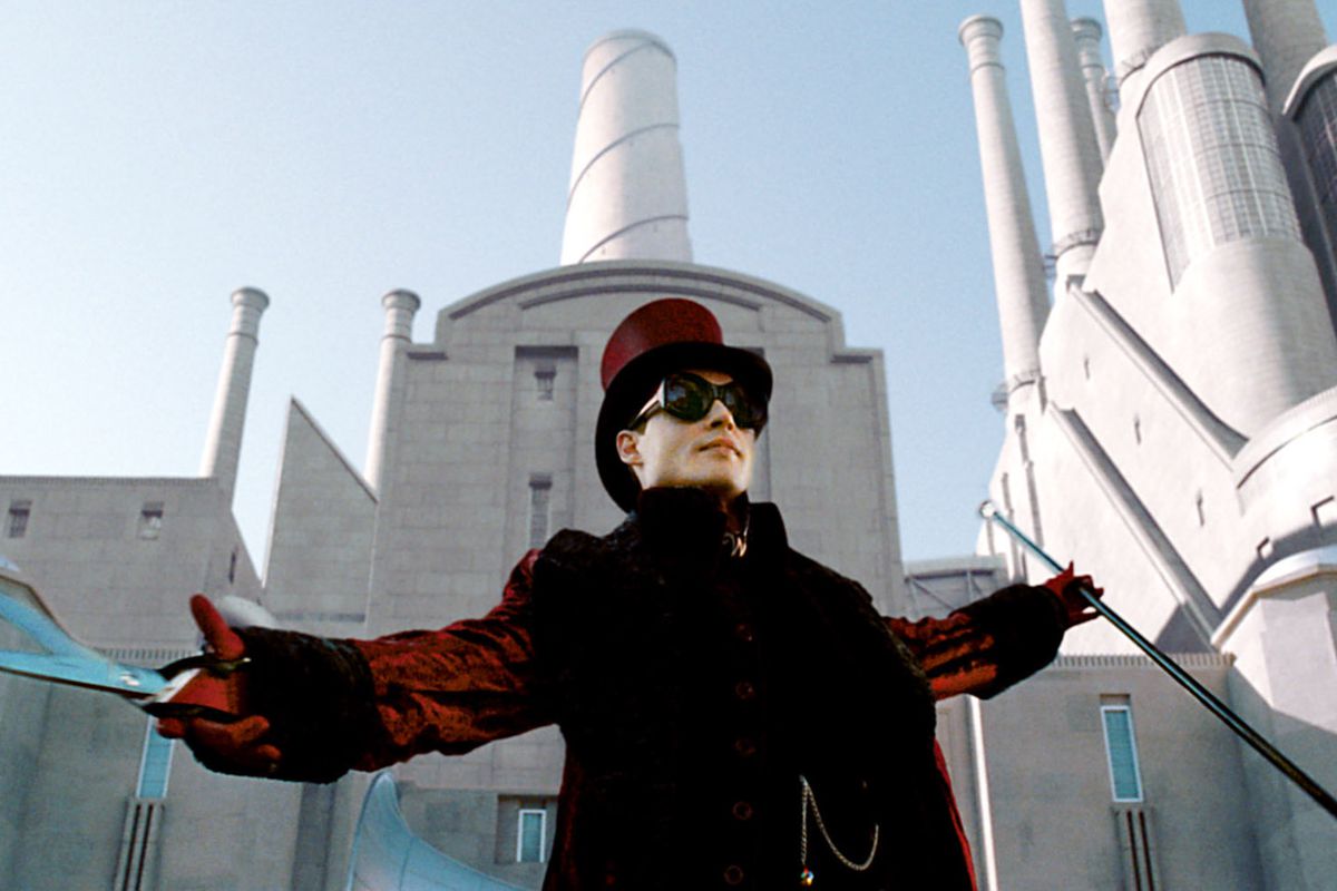 Willy Wonka (Johnny Depp, in red coat, top hat, and huge black goggles) holds up a pair of novelty sheers and stands with his arms wide in front of the all-white exterior of his chocolate factory in Tim Burton’s 2005 Charlie and the Chocolate Factory