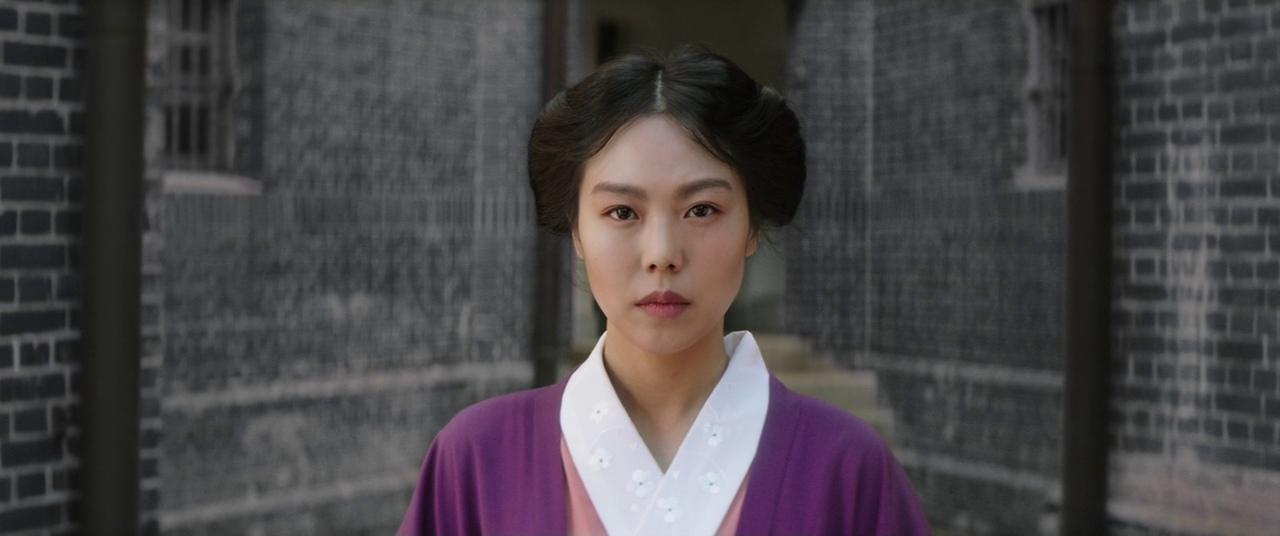 A young woman wearing a purple dress looks ahead, with dull bricks behind her, in The Handmaiden.